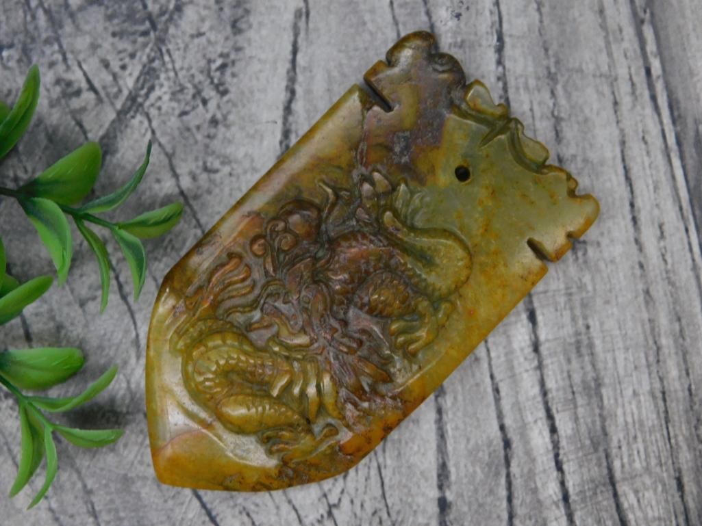 Carved Jade Pendant with Dragon Motif Rock Stone Lapidary Specimen: - A carved jade pendant featuring an intricate dragon design. | - Appears to have a hole near the top, likely for use as a pendant. | - Light brown, yellow, and green tones characteristic of natural j