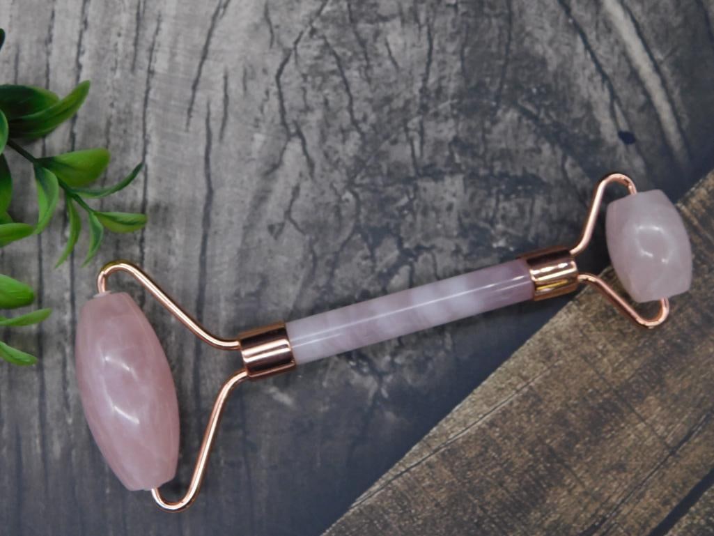 Rose Quartz Facial Roller With Copper Frame: - Rose quartz facial roller featuring two polished stone ends | - Handle made of matching rose quartz, providing a cohesive look | - Copper-plated frame holding the stones securely in place | - Dual r