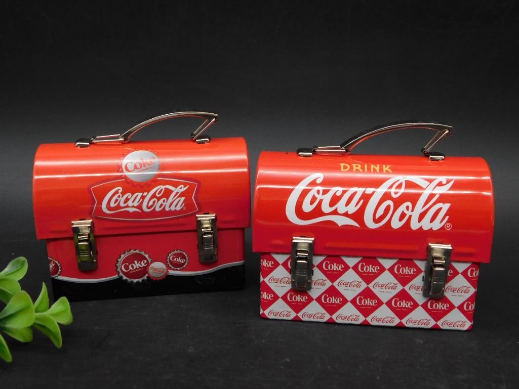 Two Coca-Cola Miniature Tin Lunch Boxes: - Set includes two miniature tin lunch boxes featuring Coca-Cola branding. | - One design features an all-red pattern with the classic Coca-Cola logo and text 'Drink Coca-Cola.' | - The other design s