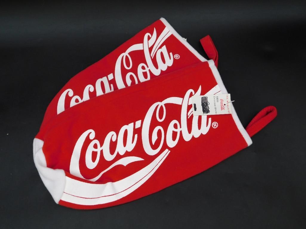 Two Coca-Cola Branded Christmas Stockings: - Set includes two Coca-Cola themed Christmas stockings. | - Both stockings are predominantly red with bold Coca-Cola logos in white. | - Comes with original tags marked 'The World of Coca-Cola' and a
