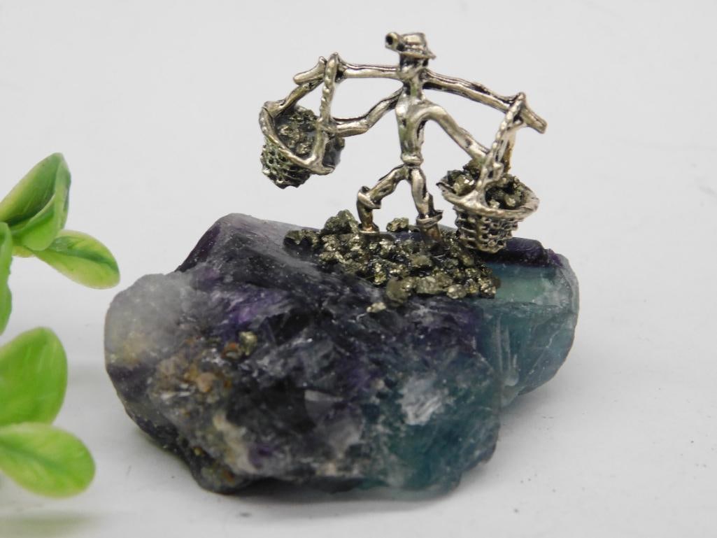 Pyrite Miner on Fluorite Base Rock Stone Lapidary Specimen: - Metal figurine depicting a person carrying two baskets, balanced on a shoulder beam. | - Mounted on a colorful crystal base featuring purples, blues, and greens. | - Metallic accents in baskets appe