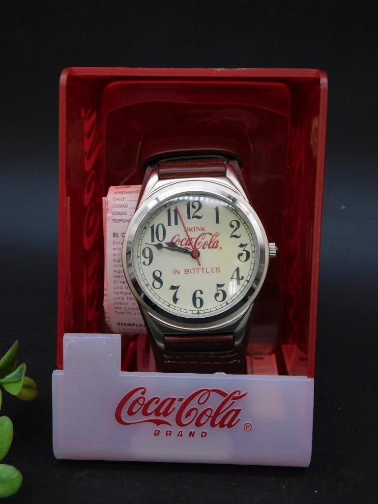 Coca-Cola Branded Watch in Original Display Case: - Coca-Cola branded wristwatch with a white dial featuring the text 'Drink Coca-Cola in Bottles.' | - Black numerals, hour markers, and metal hour, minute, and second hands. | - Stainless steel casing