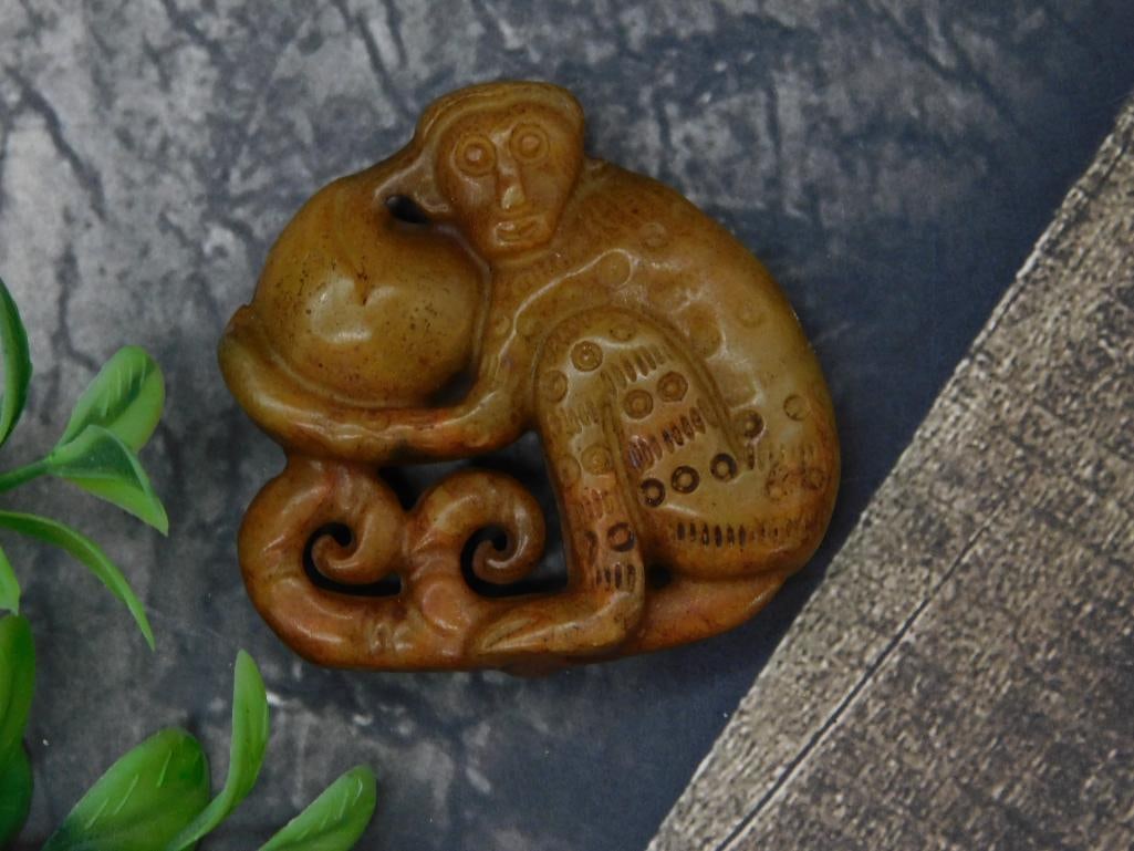 Carved Jade Stone Figurine of Monkey with Fruit: - Carved stone depicting a monkey holding a fruit. | - Brown stone material with natural variations in texture and color. | - Detailed design featuring swirling accents at the base. | - Compact size f
