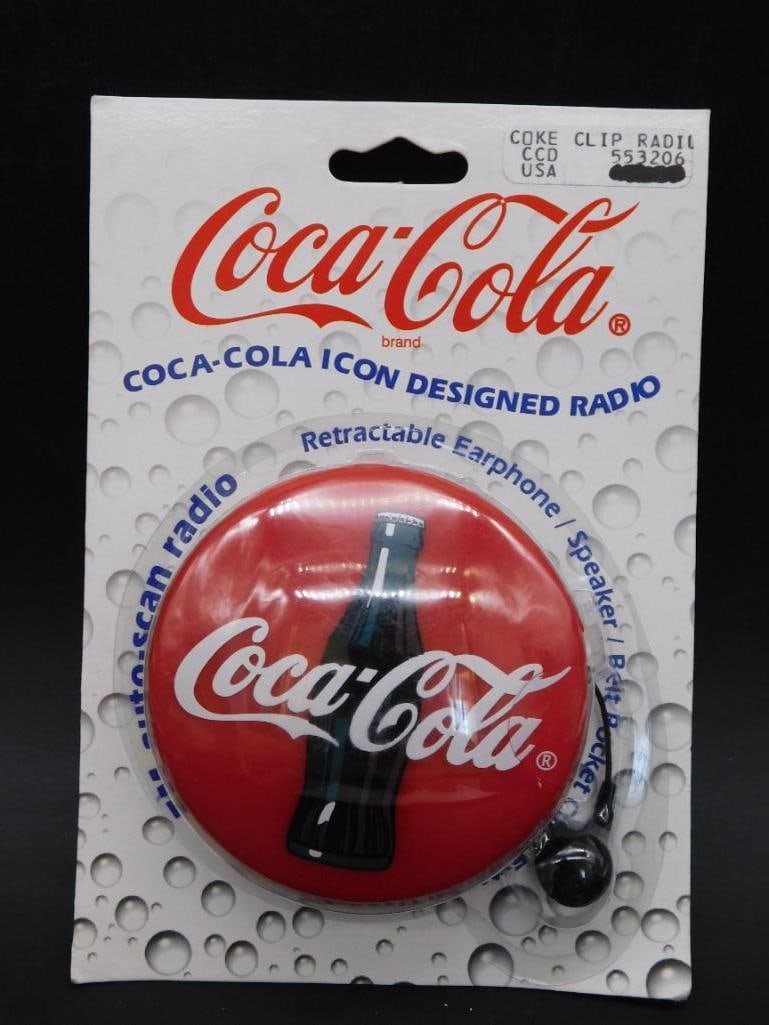 Coca-Cola Icon Designed Radio in Original Packaging: - Coca-Cola themed circular radio with brand logo and bottle design. | - Includes retractable earphone, speaker, and belt clip. | - Item is new and sealed in its original packaging. | - Appears unused