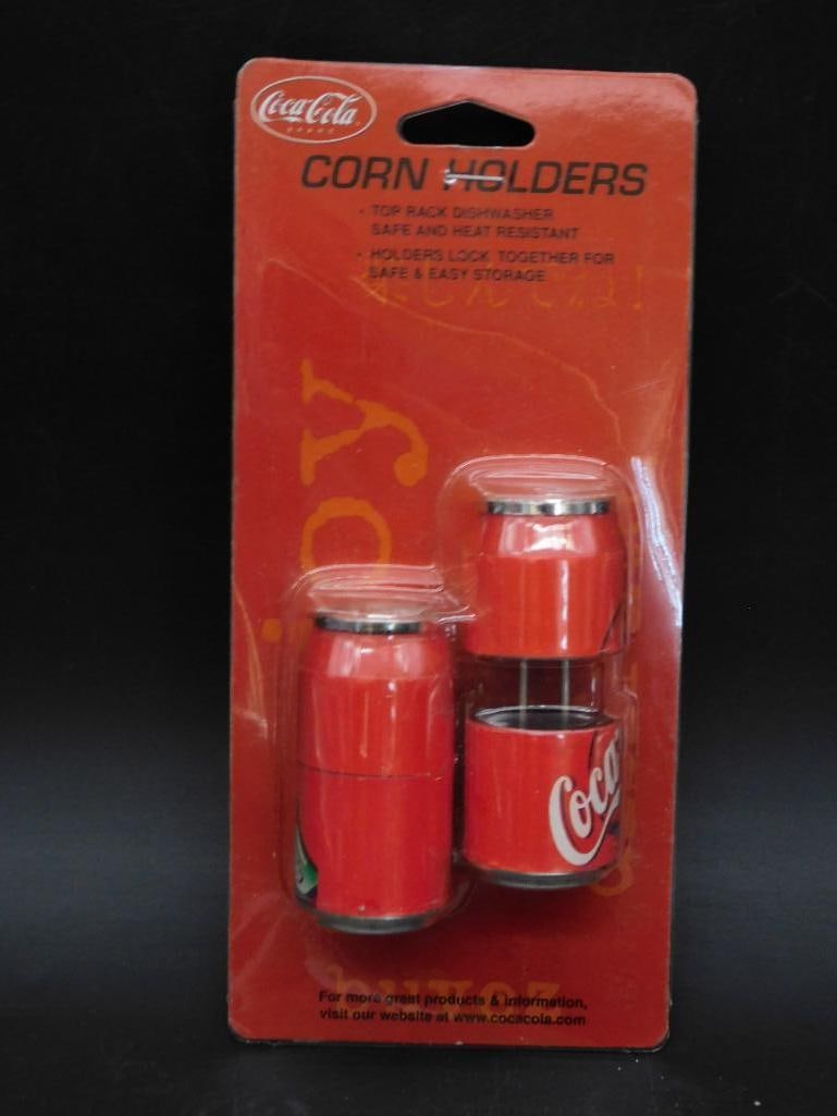 Coca-Cola Branded Corn Holders in Original Packaging: - Coca-Cola branded corn holders, set of 2 in red color. | - Comes in original sealed packaging with the Coca-Cola logo. | - Product features include: top rack dishwasher safe and heat resistant. | -