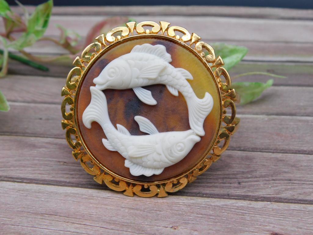 Pisces Zodiac Hinged Bracelet with Fish Design: Gold-tone hinged bracelet featuring an ornate circular design with two swimming fish in white relief, representing the Pisces zodiac sign. The backing appears to contain a marbled surface. Includes at