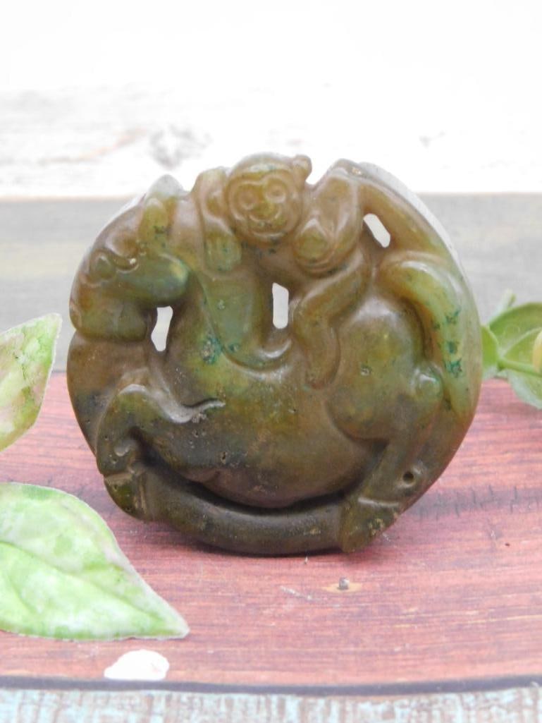 Carved Jade ROCK STONE LAPIDARY SPECIMEN: A carved green stone pendant portraying a detailed depiction of a monkey riding a horse. The piece showcases intricate craftsmanship with holes to facilitate wear or display. Auction lot information i