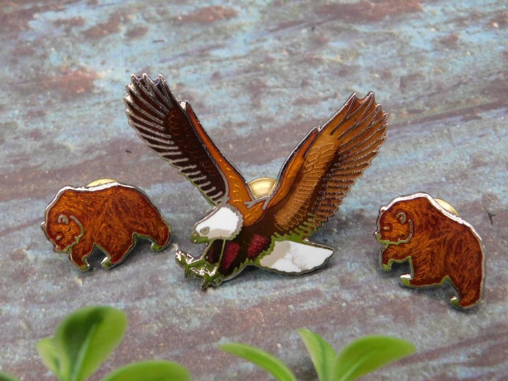 Set of Three Wildlife-Themed Metal Pins With Enamel Design: This lot includes a set of three wildlife-themed metal pins with enamel design. The pins depict a bald eagle in flight and two animals resembling bears or bison. Each pin features a vibrant enamel fin