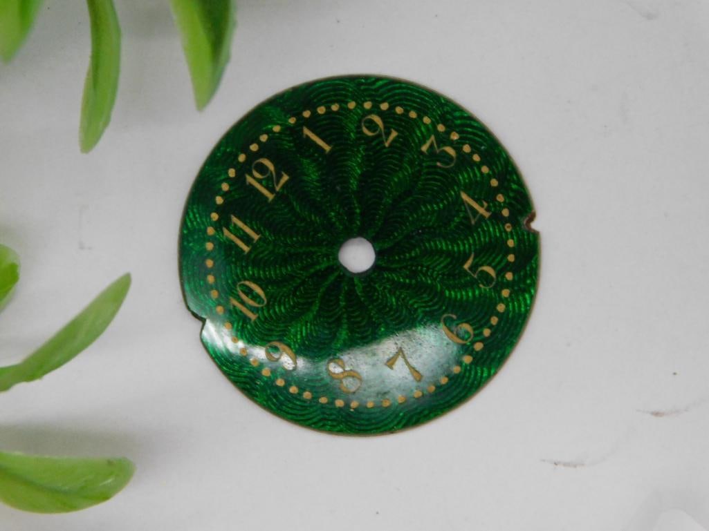 Green Enamel Watch Dial with Gold Numeric Markers WATCHMAKER WATCHMAKING TOOLS: - Green enamel watch dial with detailed guilloche pattern. | - Features gold numeric hour markers from 1 to 12. | - Circular shape with a center hole, likely for fitting watch hands. | - Sized for sma