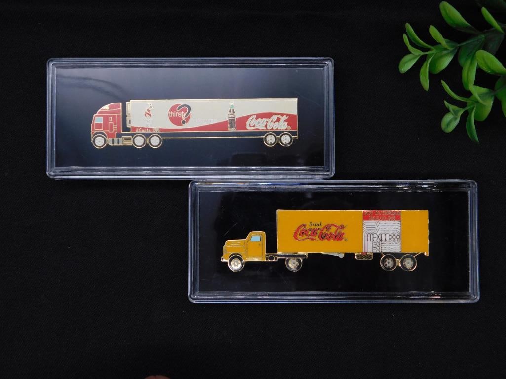 Coca-Cola Truck Lapel Pins in Plastic Cases: - Set of 2 Coca-Cola-themed truck-shaped lapel pins. | - Pins feature vibrant designs, including Coca-Cola branding and promotional graphics. | - One pin highlights the XIX Olympic Games Mexico 1968.