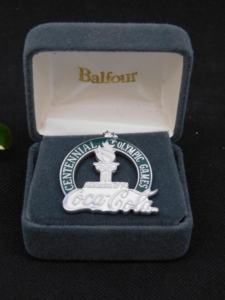 Coca-Cola Centennial Olympic Games Pin in Display Box: - Coca-Cola branded pin commemorating the Centennial Olympic Games in Atlanta, 1996. | - Features a silver-finished design with imagery of the Olympic torch. | - Includes original display box by Balfo