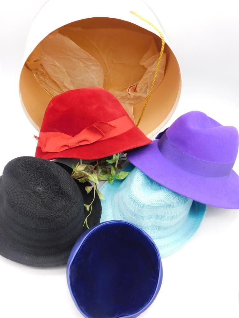 Collection of Five Hats with Hat Box: A collection of five stylish hats in different colors and designs. Includes a red hat with ribbon detail, a black hat, a purple hat, a blue straw-like hat, and a blue velvet hat. Comes with a round ha