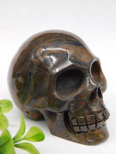 Hand-Carved Natural Stone Skull Sculpture Rock Stone Lapidary Specimen: - Features a hand-carved skull made from natural stone with visible flowing patterns. | - Striking brown hues with subtle red and green tones throughout the piece. | - Compact size, as shown with a ru