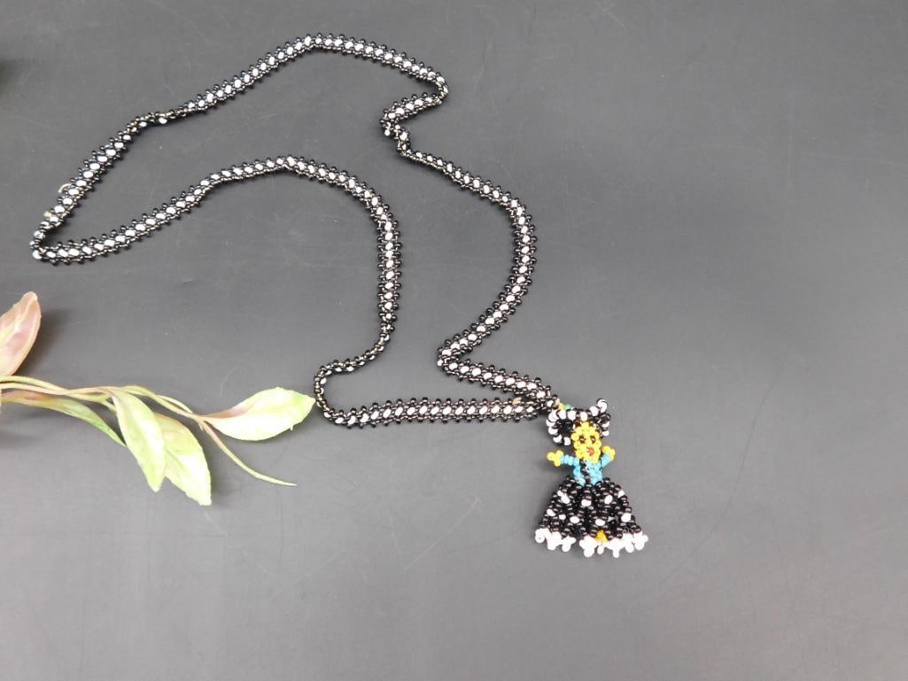 Beaded Necklace With Figurine Pendant in Black and Multicolor: Beaded necklace with a handcrafted figurine pendant. The chain features black and white beads woven together. The pendant portrays a colorful figure with a blue torso, black and white skirt, and yello