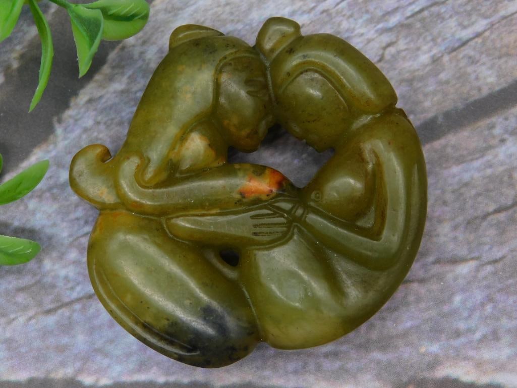 Green Jade Carving of Two Embracing Figures: A green stone carving depicting two embracing figures in a circular arrangement. The piece is crafted from a solid material with natural variations in the stone, including darker and lighter tones. In