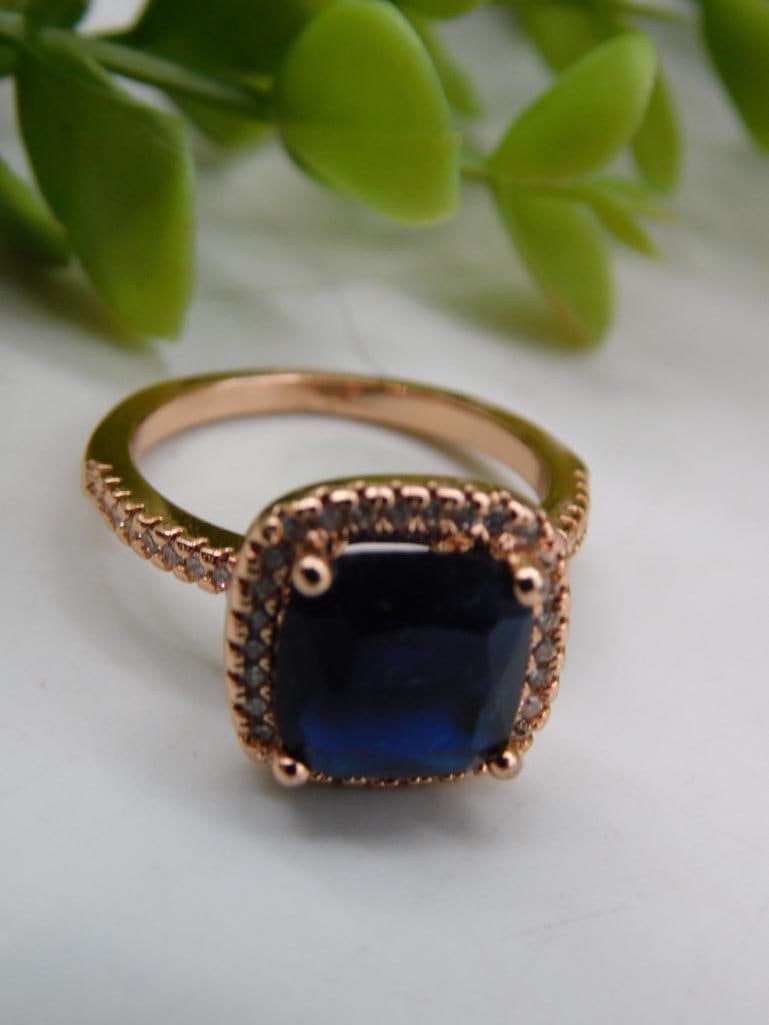 Rose Gold-Tone Ring with Sapphire-Like Center Stone: Features a rose gold-tone band with intricate detailing. | Centerpiece includes a dark blue, sapphire-like square-cut stone. | Surrounding the main stone are small clear accents enhancing the design. 