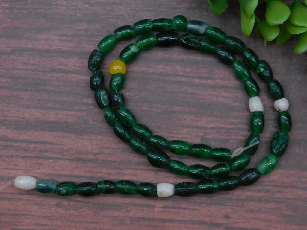 Strand of Green Glass Beads with White Accents: Strand of green glass beads with scattered white and one yellow accent bead. | Beads are oval-shaped and uniform in size. | Approximate length of strand is shown with a standard ruler for reference. |