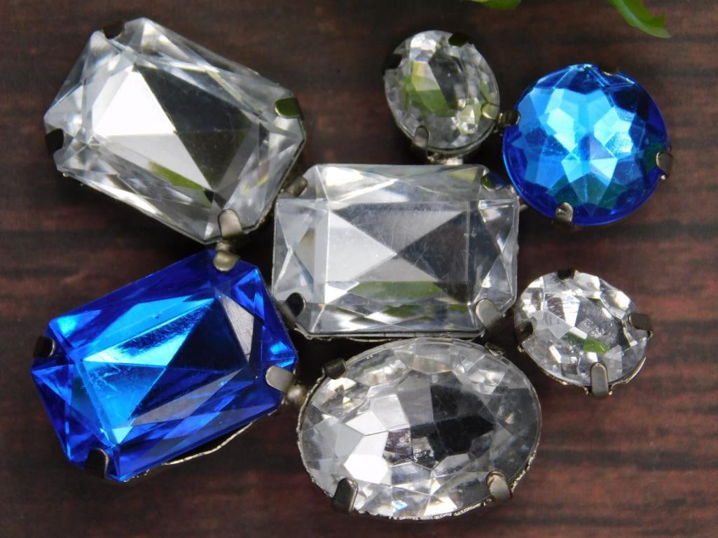 Blue and Clear Glass Rhinestones: - Mixed lot of faceted glass rhinestones. | - Includes both blue and clear stones in various shapes and sizes. | - Mounted in metal prong settings for easy use. | - Displayed next to a quarter for siz
