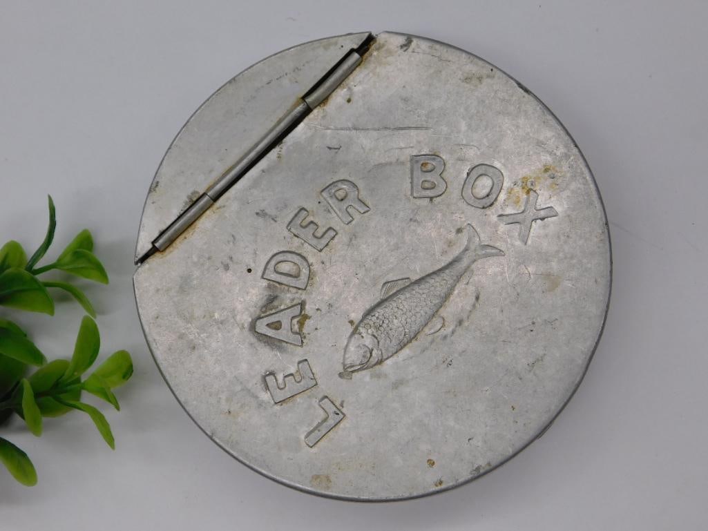 Vintage Metal Leader Box with Embossed Fish Design: - Round metal box marked 'Leader Box' with an embossed fish design. | - Made of metal and features a hinged lid for secure closure. | - Appears to contain an unidentified contents resembling material