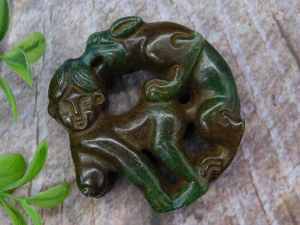 Carved Jade Stone Figurine With Animal Motif: This item is a carved green stone figurine depicting a human face intertwined with an animal, possibly a rabbit. The intricate design features flowing curves and detailed carving work, showcasing a dy