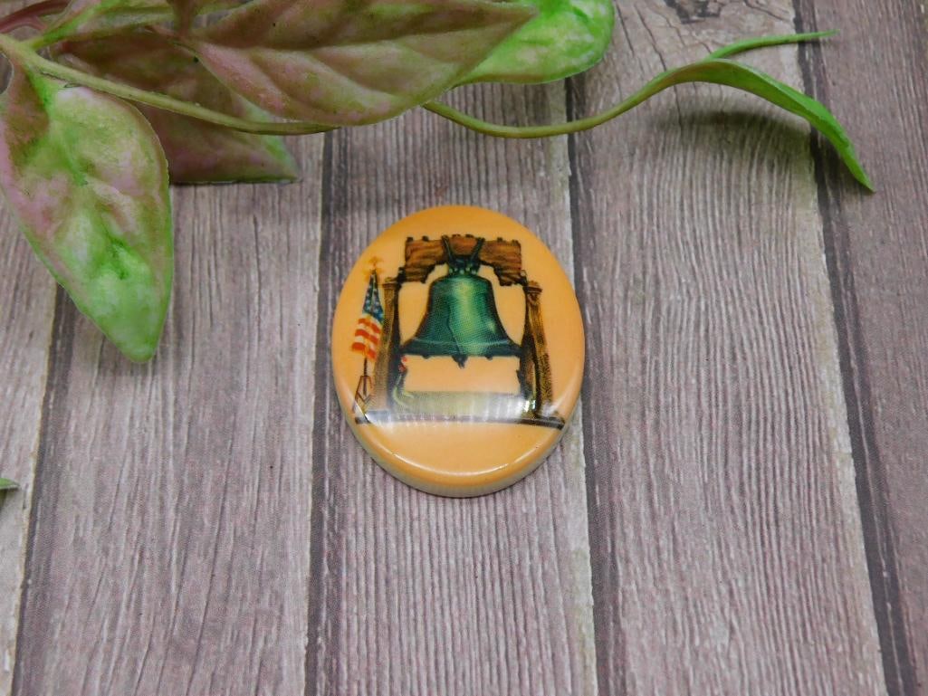 Circular Pin Featuring Liberty Bell and U.S. Flag Design: Circular pin with a design showcasing the Liberty Bell and U.S. flag. Features a yellow background with green and blue tones on the Bell. Perfectly round with a smooth surface and pinback closure. Mea