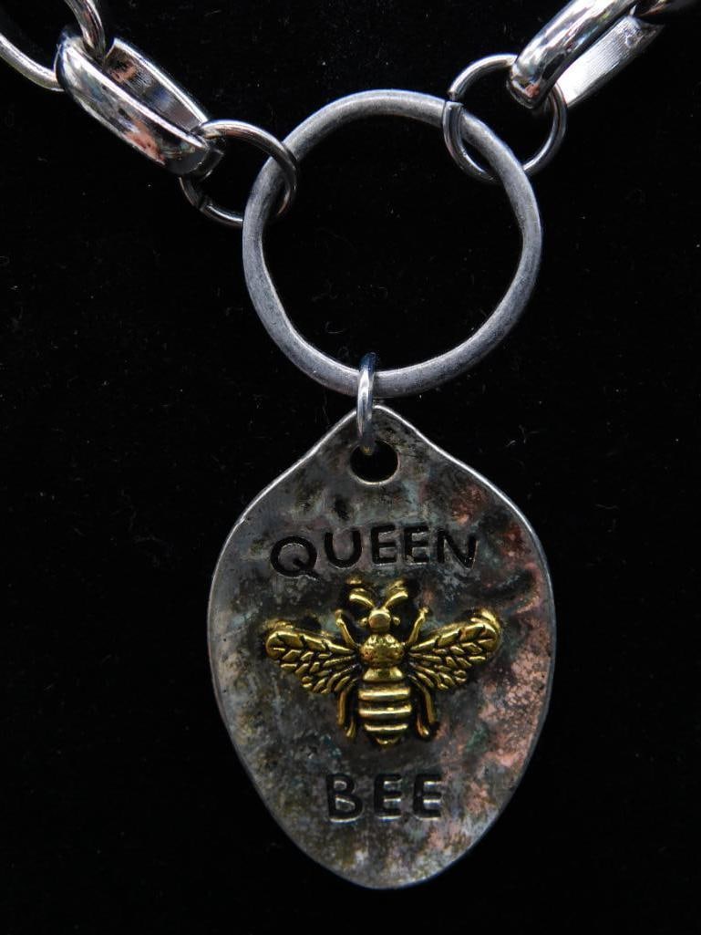 Metal Chain Bracelet with ?Queen Bee? Pendant: A sturdy metal chain bracelet featuring an oval-shaped charm with the words 'Queen Bee' engraved. The pendant showcases a three-dimensional bee motif in gold-tone against the textured backgrou
