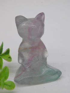 Carved Fluorite Cat Figurine ROCK STONE LAPIDARY SPECIMEN: This lot features a carved fluorite cat figurine with subtle hues of purple, green, and pink. The figurine showcases a seated cat atop a flat base. The material displays natural banding and transparen