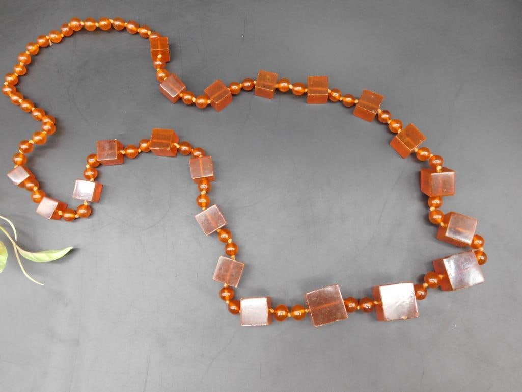 Amber-Colored Beaded Necklace With Square Accents: Amber-colored beaded necklace featuring alternating round and square-shaped beads. The square beads have a polished surface, while the round beads are smaller and uniformly shaped. The necklace is str
