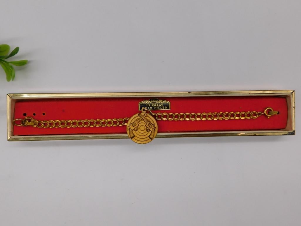 12 Karat Gold-Filled Shooting Medal Bracelet: - Features a 12 karat gold-filled construction, as indicated on the tag. | - Includes a medal charm with crossed pistols and a target design. | - Chain bracelet with a secure clasp closure. | - Origin