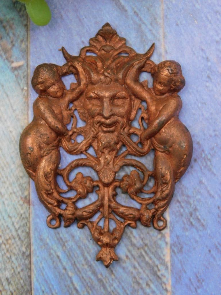 Antique Cast Iron Decorative Plaque with Mythological Motif: Cast iron decorative plaque featuring a mythological face and two cherub-like figures. | Intricate openwork design with detailed ornamentation. | Appears to have a bronze or copper-toned finish. | Des