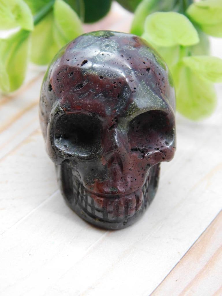 Stone Skull Figurine with Dark Red and Green Hues: Hand-carved stone skull figurine featuring dark red and green accents. | Displayed on a wooden surface with a quarter nearby for size reference. | Lightweight and compact; a suitable collectible or de