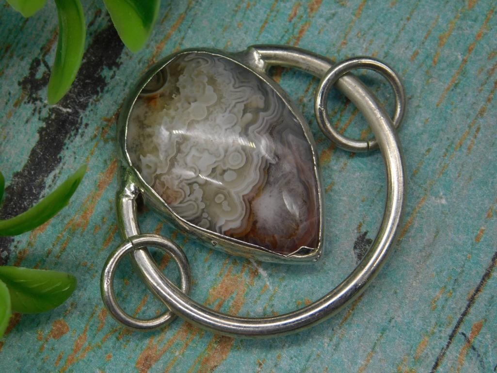 CRAZY LACE Pendant with Teardrop-Shaped Banded Agate Stone: Unique pendant featuring a teardrop-shaped banded agate stone. The stone displays striking natural patterns and earth tones. Encased in a silver-tone metal setting with decorative looped accents. Incl
