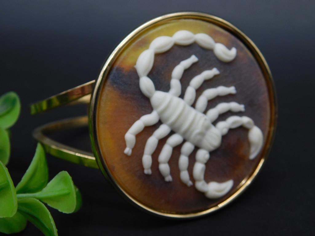 Gold-Plated Bracelet With Scorpion Cameo Design: This gold-plated bracelet features a prominent scorpion cameo design. The bracelet has an oval face with a detailed scorpion figure in white against a brown background. The hinge mechanism provides