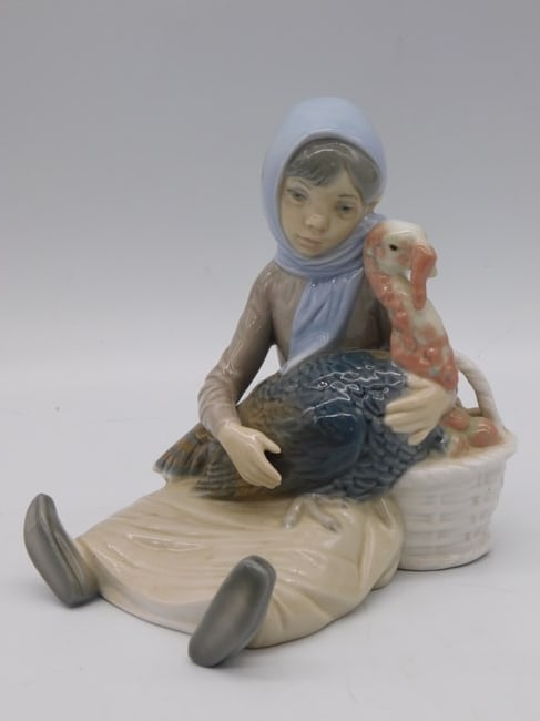 Lladro Style Porcelain Figurine of Woman with Turkey and Basket: A porcelain figurine depicting a woman in a blue headscarf carrying a large turkey under one arm and a woven basket over the other. The piece is marked on the base LLADRO Hand Made in Spain with an im