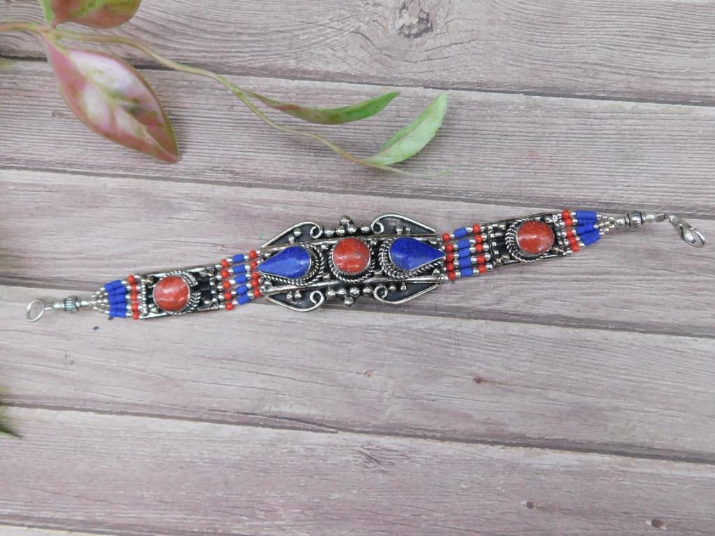 Lapis Lazuli and Red Coral Bracelet: This bracelet features a silver-tone base with ornate detailing, accented by red and blue beads. The design has a clasp closure, making it functional and secure for wear. The bracelet?s size aligns