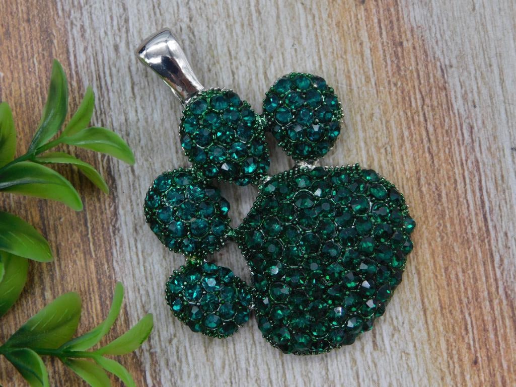 Green Rhinestone Paw Print Pendant: A silver-tone pendant shaped like an animal paw print, fully set with sparkling green rhinestones. A fun, wearable piece for pet lovers and costume jewelry fans. Shown next to a quarter for scale.