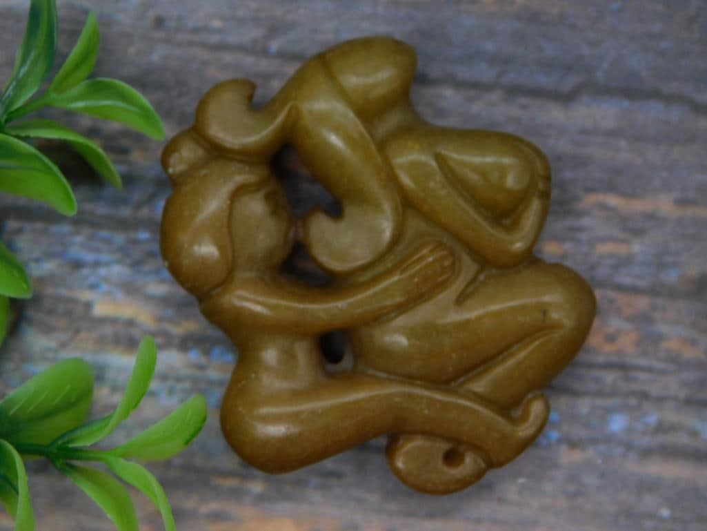 Carved Stone Figural Pendant - Abstract Jade-Tone Amulet: A hand-carved stone pendant in an abstract figural design, finished in a warm olive-brown tone with a drilled hanging hole. The pendant measures approximately 2 inches across. An interesting