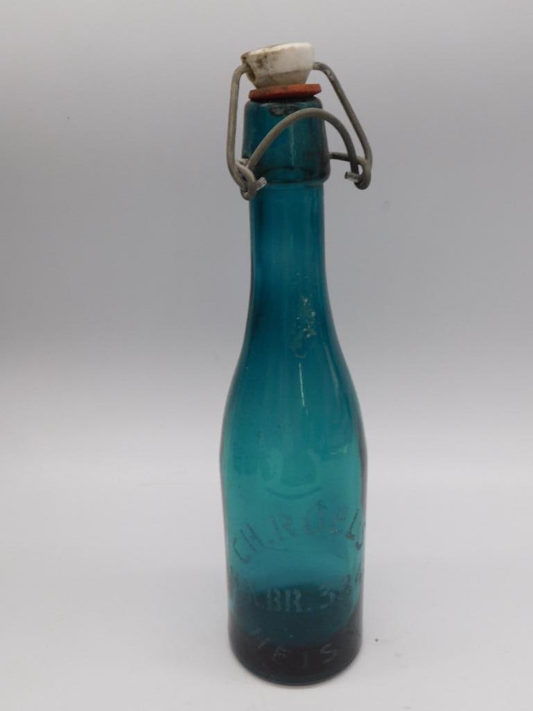 Antique Aqua Glass Bottle with Wire Bail Stopper: An antique aqua blue glass bottle with embossed lettering and its original porcelain and wire bail swing top stopper. Stands approximately 11 inches tall. A nice piece for antique bottle collectors.