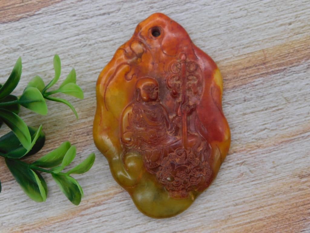 Carved Chinese Soapstone Pendant with Deity Figure, Orange & Green: A hand-carved soapstone pendant in mottled orange, red, and green hues, depicting a seated deity figure amid scrollwork with a drilled hanging hole. Measures approximately 3 inches long by 2 inches