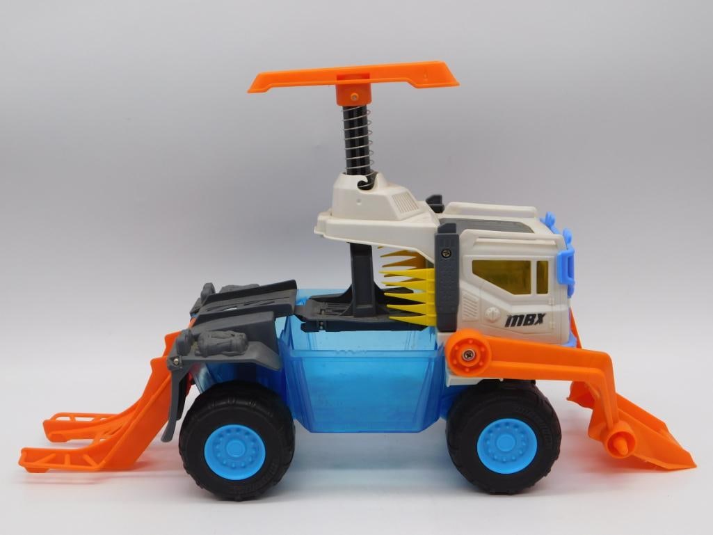 Matchbox MBX Off-Road Construction Truck Toy with Plow & Scoop: A Matchbox MBX-branded off-road construction vehicle in blue, orange, and white with a translucent blue cargo bed, front snow plow, rear scoop arm, spring-loaded roll bar, and rugged tires. Marked Mat