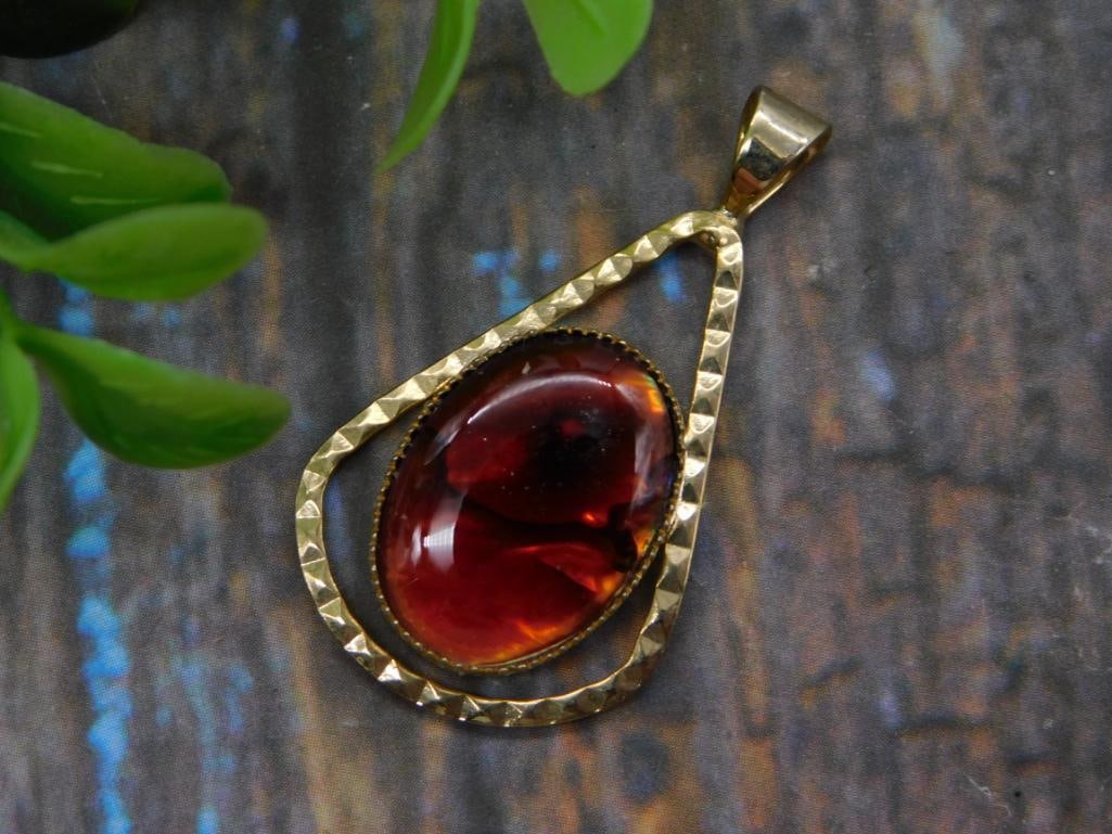 Amber/Garnet-Style Oval Cabochon Pendant, Gold-Tone Teardrop Setting: A deep red-amber oval glass or resin cabochon pendant set within a hammered gold-tone teardrop frame with a decorative bail. Measures approximately 2 inches long, shown with a 1966 quarter for scale; 