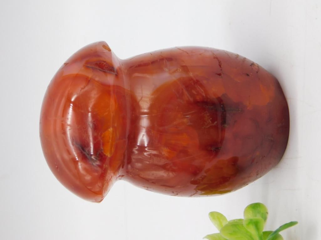 Carved Carnelian Agate Stone Figure, Polished Red-Orange: Polished carnelian or banded agate carved into a stylized hourglass/vase form with rich translucent red-orange coloring and natural veining. Measures approximately 4 inches long, shown beside a