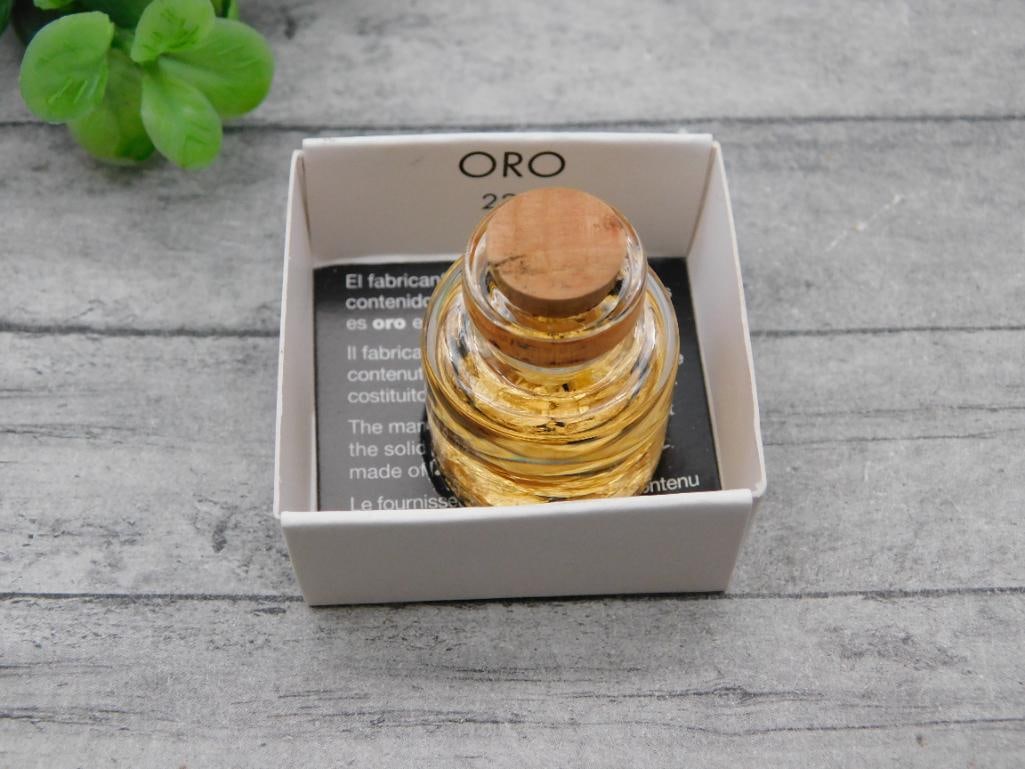 Antique Gold Flake Novelty Bottle: Small corked glass bottle labeled ORO containing floating gold leaf flakes suspended in liquid, presented in its original small cardboard display box with multilingual text. Shown with a quarter for