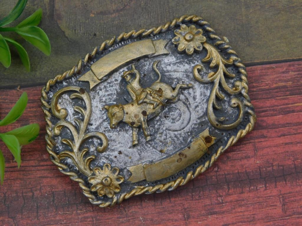 Western Rodeo Belt Buckle: A tarnished silver-and-gold tone metal belt buckle featuring an embossed bull rider scene framed with rope border and floral scroll accents. Blank banners for engraving