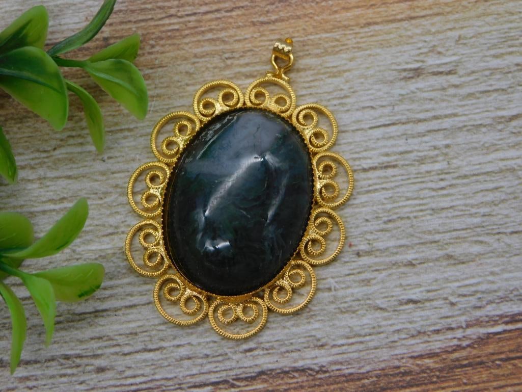 Moss Agate Gold Filigree Pendant: An oval moss agate cabochon pendant in deep green-black tones set in an ornate gold-tone filigree scrollwork frame. Shown with a quarter for scale, roughly 2 inches long.