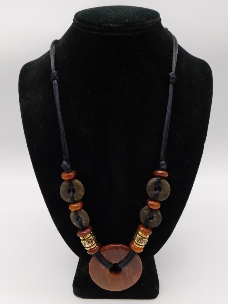 Tribal Style Beaded Stone Necklace: A handmade necklace featuring a large round carnelian/agate pendant disc with wood, brass, and orange bead accents on a black cord. Approximately 24 inches long.