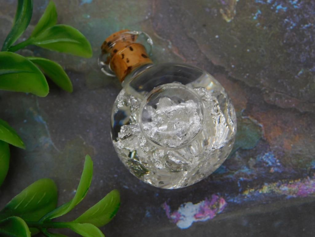 Crushed Quartz Crystal Glass Bottle: A small round glass bottle with cork stopper filled with crushed clear quartz crystal chips.