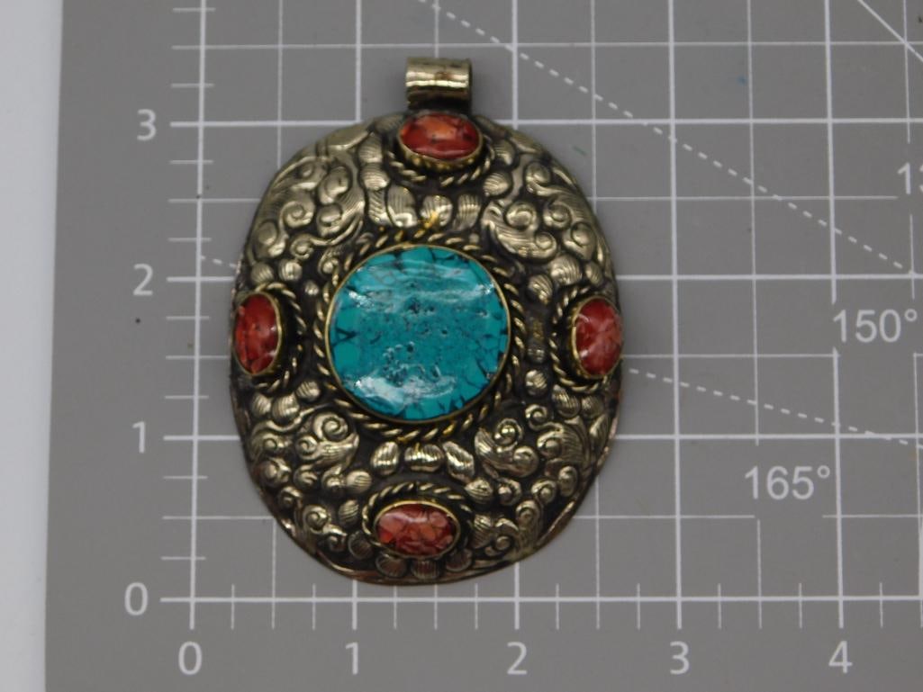 Jewelry Tibetan Silver Turquoise and Coral Pendant: An oval silver-tone pendant with repousse scrollwork surrounding a central round turquoise cabochon and four smaller coral cabochon accents, fitted with a tube bail for stringing. Measures