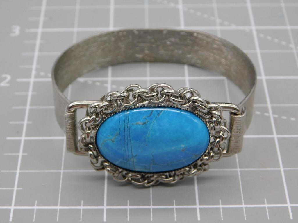 Turquoise Cabochon Bangle Bracelet: A silver-tone hinged bangle bracelet centered with a large oval turquoise-colored cabochon set within a decorative rope-chain bezel. The bracelet has a brushed metal band and measures roughly 3