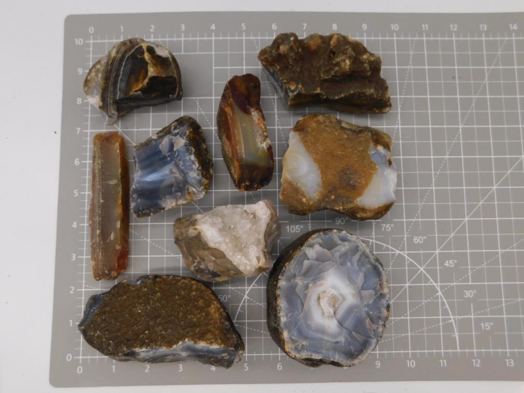 Agate Geode Rough Specimen Lot: Nine rough agate and geode specimens with banded blue-gray, white, and brown chalcedony faces, several with visible crystal-lined cavities, shown on a ruled cutting mat. Total weight labeled 5 lbs 7
